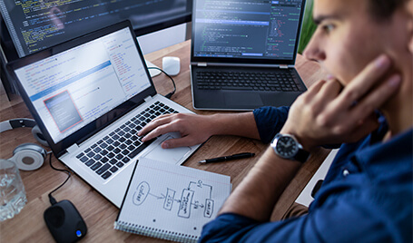 A man is sitting at a table and typing something on a laptop. There is a notepad, computer mouse, headphones, and a second laptop next to him. A PC monitor screen is visible in the background.