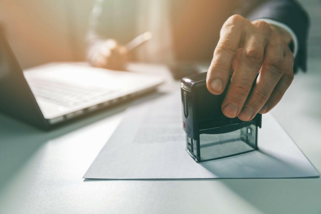 A man, sitting at a desk with a laptop on it, is placing a seal on a document.